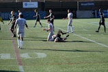 A soccer match scene where one player is on the ground while others look on. The players are wearing different colored uniforms, indicating opposing teams. The field is made of artificial turf with visible lines.