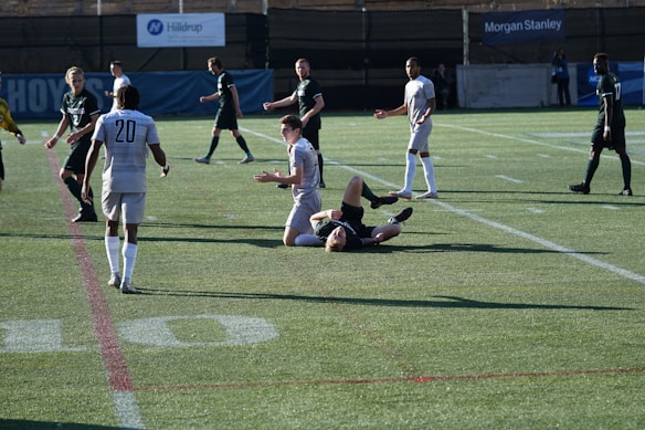 A soccer match scene where one player is on the ground while others look on. The players are wearing different colored uniforms, indicating opposing teams. The field is made of artificial turf with visible lines.