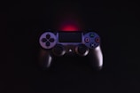 A video game controller with a dark finish rests against a black background. The controller features the classic directional pad and buttons highlighted with blue, pink, and green symbols. A subtle red backlight creates a glow above the controller.