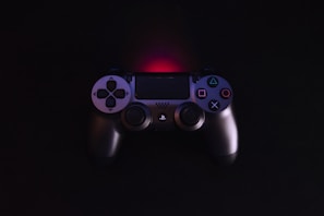 A video game controller with a dark finish rests against a black background. The controller features the classic directional pad and buttons highlighted with blue, pink, and green symbols. A subtle red backlight creates a glow above the controller.