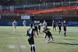 A group of male soccer players are engaged in a competitive match on a grass pitch. The players are wearing jerseys with numbers and are in motion, with one player attempting to head a soccer ball. The scene includes daylight and a background with banners and a low fence. Spectators or security personnel can be seen in the distance on an elevated walkway.