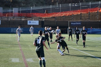 A group of male soccer players are engaged in a competitive match on a grass pitch. The players are wearing jerseys with numbers and are in motion, with one player attempting to head a soccer ball. The scene includes daylight and a background with banners and a low fence. Spectators or security personnel can be seen in the distance on an elevated walkway.