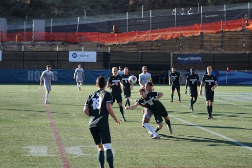 A group of male soccer players are engaged in a competitive match on a grass pitch. The players are wearing jerseys with numbers and are in motion, with one player attempting to head a soccer ball. The scene includes daylight and a background with banners and a low fence. Spectators or security personnel can be seen in the distance on an elevated walkway.