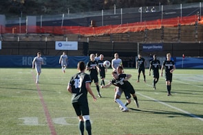 A group of male soccer players are engaged in a competitive match on a grass pitch. The players are wearing jerseys with numbers and are in motion, with one player attempting to head a soccer ball. The scene includes daylight and a background with banners and a low fence. Spectators or security personnel can be seen in the distance on an elevated walkway.
