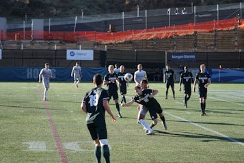 A group of male soccer players are engaged in a competitive match on a grass pitch. The players are wearing jerseys with numbers and are in motion, with one player attempting to head a soccer ball. The scene includes daylight and a background with banners and a low fence. Spectators or security personnel can be seen in the distance on an elevated walkway.