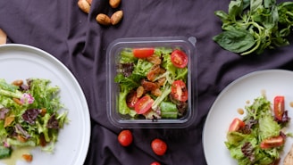 A selection of fresh salads is displayed on a dark cloth. Two white plates contain mixed greens, cherry tomatoes, and nuts, with a central plastic container featuring a similar salad. A handful of whole almonds are scattered around along with loose cherry tomatoes. Fresh basil leaves are placed in the top right corner.