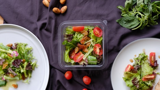 A selection of fresh salads is displayed on a dark cloth. Two white plates contain mixed greens, cherry tomatoes, and nuts, with a central plastic container featuring a similar salad. A handful of whole almonds are scattered around along with loose cherry tomatoes. Fresh basil leaves are placed in the top right corner.