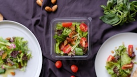 A selection of fresh salads is displayed on a dark cloth. Two white plates contain mixed greens, cherry tomatoes, and nuts, with a central plastic container featuring a similar salad. A handful of whole almonds are scattered around along with loose cherry tomatoes. Fresh basil leaves are placed in the top right corner.