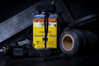 A bottle of USMC black leather dye is at the center, surrounded by various crafting tools. The label is prominent, featuring bold yellow with black text and warning information. Nearby are spools of dark thread and a tool with a textured handle, all set on a dark surface with low lighting.