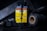 A bottle of USMC black leather dye is at the center, surrounded by various crafting tools. The label is prominent, featuring bold yellow with black text and warning information. Nearby are spools of dark thread and a tool with a textured handle, all set on a dark surface with low lighting.