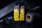 A bottle of USMC black leather dye is at the center, surrounded by various crafting tools. The label is prominent, featuring bold yellow with black text and warning information. Nearby are spools of dark thread and a tool with a textured handle, all set on a dark surface with low lighting.