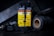 A bottle of USMC black leather dye is at the center, surrounded by various crafting tools. The label is prominent, featuring bold yellow with black text and warning information. Nearby are spools of dark thread and a tool with a textured handle, all set on a dark surface with low lighting.