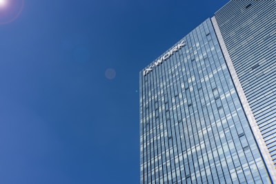A tall modern skyscraper with reflective glass windows dominates the right side of the image, with a clear blue sky filling the rest. The building&rsquo;s facade is sleek and streamlined, featuring horizontal stripes of glass. The name 'IX WORK' is visible at the top of the building.