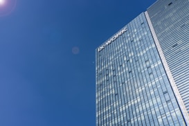 A tall modern skyscraper with reflective glass windows dominates the right side of the image, with a clear blue sky filling the rest. The building&rsquo;s facade is sleek and streamlined, featuring horizontal stripes of glass. The name 'IX WORK' is visible at the top of the building.