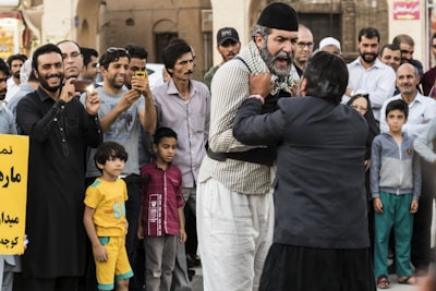 A crowded street scene with a group of people gathered around two men in the center engaged in what seems to be a heated argument or performance. Many onlookers are smiling or filming with their phones. The crowd includes men, women, and children, all dressed in casual clothes. There is a sign in the foreground with text in a non-Latin script.