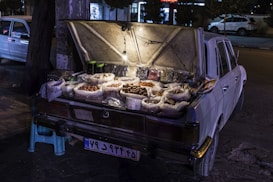 An old car is parked with its trunk open, used as a makeshift shop to sell various types of nuts and dried fruits. The trunk is illuminated by a single light bulb hanging from the open lid, showcasing multiple plastic bags filled with assorted goods. The car is parked on the street at night, with a stool placed beside it.
