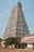 A tall, intricately carved temple tower with vibrant colors and detailed sculptures, typical of South Indian architecture. The scene includes a clear sky and a tree in the foreground, along with some structures surrounding the temple.