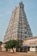 A tall, intricately carved temple tower with vibrant colors and detailed sculptures, typical of South Indian architecture. The scene includes a clear sky and a tree in the foreground, along with some structures surrounding the temple.