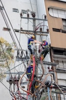 Two electrical workers in safety gear are working high up on a utility pole surrounded by numerous wires and equipment. They are in a cityscape environment, and the background shows buildings with windows. The workers are using tools to handle the complex array of wires.