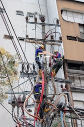 Two electrical workers in safety gear are working high up on a utility pole surrounded by numerous wires and equipment. They are in a cityscape environment, and the background shows buildings with windows. The workers are using tools to handle the complex array of wires.