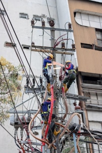 Two electrical workers in safety gear are working high up on a utility pole surrounded by numerous wires and equipment. They are in a cityscape environment, and the background shows buildings with windows. The workers are using tools to handle the complex array of wires.