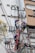 Two electrical workers in safety gear are working high up on a utility pole surrounded by numerous wires and equipment. They are in a cityscape environment, and the background shows buildings with windows. The workers are using tools to handle the complex array of wires.
