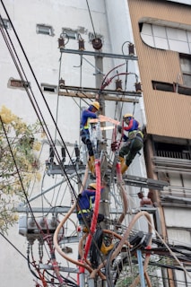 Two electrical workers in safety gear are working high up on a utility pole surrounded by numerous wires and equipment. They are in a cityscape environment, and the background shows buildings with windows. The workers are using tools to handle the complex array of wires.