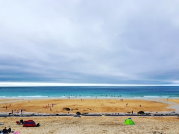 A wide sandy beach with several people walking and playing, while a few are in the water near the shore. The sea is calm with gentle waves, and the sky is overcast with clouds. Two tents, one red and one green, are set up on the sand.