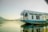 A traditional houseboat with white wooden walls and blue base reflects on the calm water. The structure has intricately carved arches and a flat roof, providing a serene and picturesque setting. The background features distant hills under a soft, hazy sky.