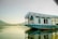 A traditional houseboat with white wooden walls and blue base reflects on the calm water. The structure has intricately carved arches and a flat roof, providing a serene and picturesque setting. The background features distant hills under a soft, hazy sky.