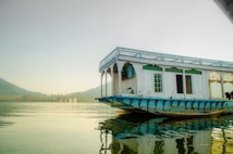 A traditional houseboat with white wooden walls and blue base reflects on the calm water. The structure has intricately carved arches and a flat roof, providing a serene and picturesque setting. The background features distant hills under a soft, hazy sky.