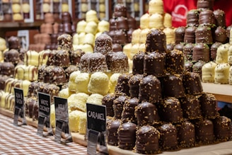 Several stacks of assorted chocolate-covered confections are displayed on a checkered tablecloth. The treats vary in flavors, indicated by small signs labeled gingerbread, white coconut, milky coconut, and caramel. Each stack features chocolates drizzled or sprinkled with toppings, creating an appealing and abundant display of sweets.
