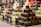 Several stacks of assorted chocolate-covered confections are displayed on a checkered tablecloth. The treats vary in flavors, indicated by small signs labeled gingerbread, white coconut, milky coconut, and caramel. Each stack features chocolates drizzled or sprinkled with toppings, creating an appealing and abundant display of sweets.