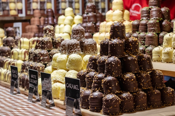 Several stacks of assorted chocolate-covered confections are displayed on a checkered tablecloth. The treats vary in flavors, indicated by small signs labeled gingerbread, white coconut, milky coconut, and caramel. Each stack features chocolates drizzled or sprinkled with toppings, creating an appealing and abundant display of sweets.