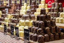 Several stacks of assorted chocolate-covered confections are displayed on a checkered tablecloth. The treats vary in flavors, indicated by small signs labeled gingerbread, white coconut, milky coconut, and caramel. Each stack features chocolates drizzled or sprinkled with toppings, creating an appealing and abundant display of sweets.