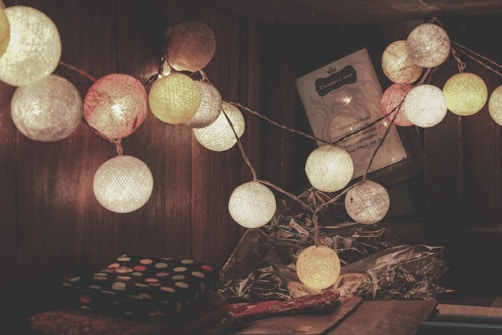 A string of spherical lights in various colors serves as decoration, illuminating a warm-toned, wooden background. The lights are wrapped in a textured string, giving a cozy atmosphere. Beneath them is a surface with fabric or paper, and a framed picture or sign is partially visible.