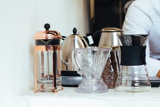 A collection of coffee-making equipment is arranged on a white surface. The setup includes a copper-colored French press, a glass pitcher, a metallic kettle, a jar filled with coffee beans, and a clear coffee dripper. A person wearing a white shirt with small dark patterns is partially seen in the background.