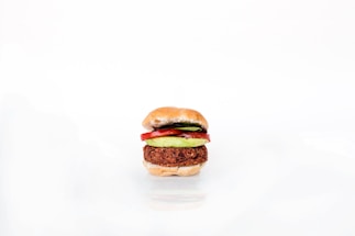 A neatly assembled vegetarian burger featuring a patty, avocado slices, and tomato slices, all sandwiched between a lightly toasted bun. The image is minimalistic with a white background highlighting the burger's colorful ingredients.