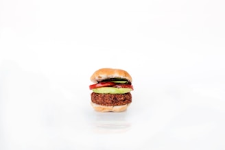 A neatly assembled vegetarian burger featuring a patty, avocado slices, and tomato slices, all sandwiched between a lightly toasted bun. The image is minimalistic with a white background highlighting the burger's colorful ingredients.
