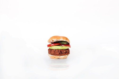 A neatly assembled vegetarian burger featuring a patty, avocado slices, and tomato slices, all sandwiched between a lightly toasted bun. The image is minimalistic with a white background highlighting the burger's colorful ingredients.