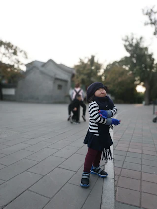 A young child dressed warmly in a striped top, gloves, and a hat stands on a paved area, gazing upward. The background features blurred images of another person, some trees, and a building with a traditional design.
