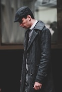 A man wearing a dark, textured overcoat and a matching newsboy cap walks with his head slightly bowed. The attire appears vintage, hinting at an old-fashioned or classic style. The background is blurred, focusing attention on the man's thoughtful expression.