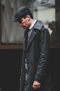 A man wearing a dark, textured overcoat and a matching newsboy cap walks with his head slightly bowed. The attire appears vintage, hinting at an old-fashioned or classic style. The background is blurred, focusing attention on the man's thoughtful expression.