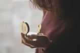 A person wearing a maroon sweater is holding an open brass compass, partially visible, with light reflecting off it. The background is softly blurred, creating a serene ambiance.