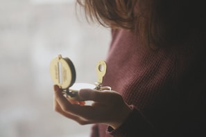 A person wearing a maroon sweater is holding an open brass compass, partially visible, with light reflecting off it. The background is softly blurred, creating a serene ambiance.