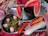 A tray contains several food items: sliced watermelon, two whole carrots, a peeled cucumber, and a side dish in a small container featuring chopped green chilies, coriander leaves, and sliced red onions. The tray is placed on a colorful fabric with geometric and floral patterns.