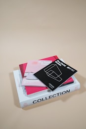 A neatly stacked assortment of books and brochures on a neutral beige surface. The top item is a black booklet titled 'The Field Guide' with a minimalist line drawing on the cover. Below it is a pink booklet and a heavier white book titled 'COLLECTION'. The arrangement is precise, creating a clean and organized aesthetic.