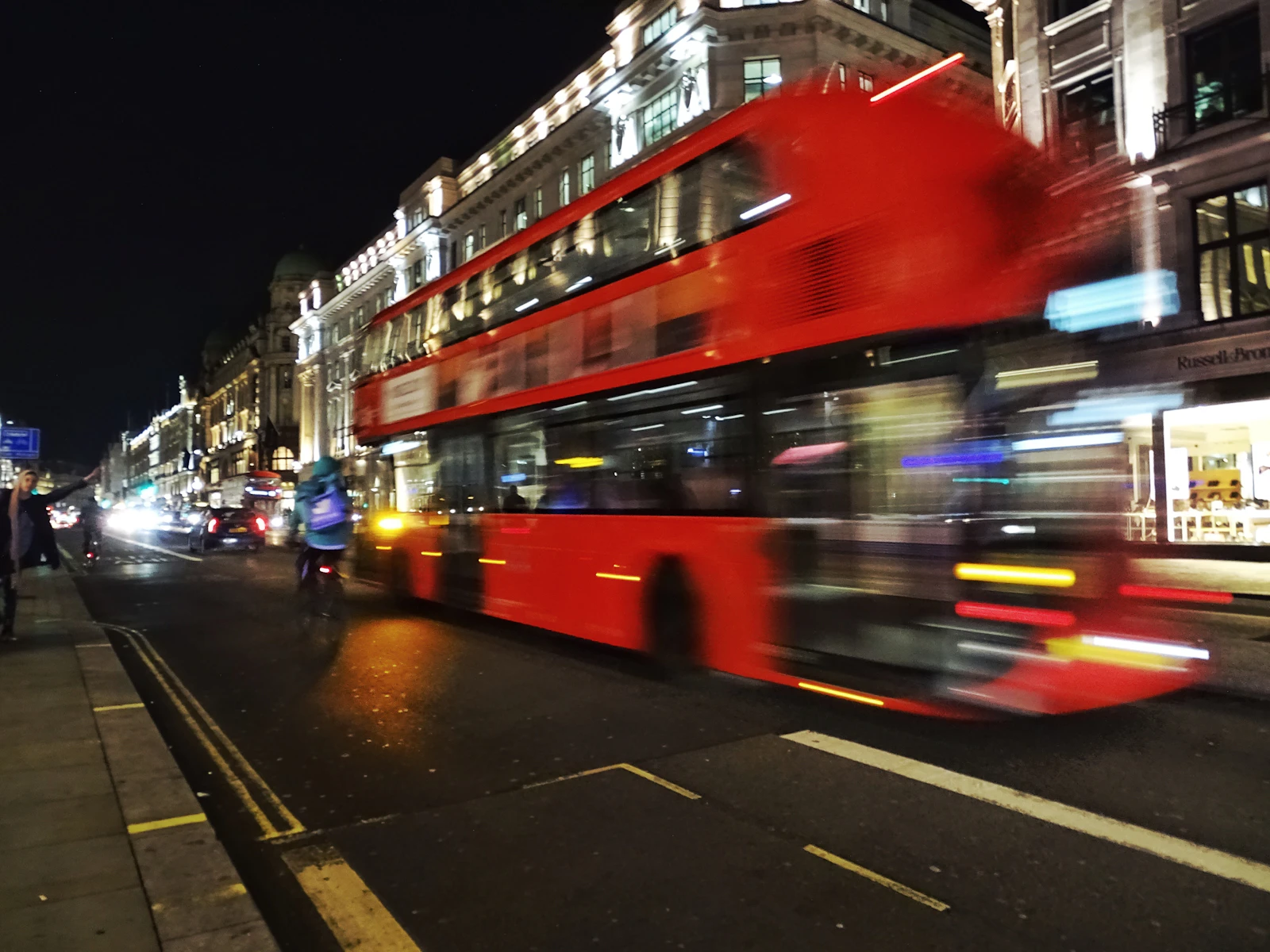 Photograph of A red double-decker bus blurs past illuminated buildings on a London street at night.