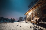 A rustic stone cabin is surrounded by snow under a clear, starry sky. The warm glow from the cabin lights contrasts with the cold blue of the night. A pair of skis and poles are propped against the building, and a bench with a blanket is placed outside, indicating a cozy yet adventurous atmosphere. Snow-covered trees are visible in the background.