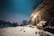 A rustic stone cabin is surrounded by snow under a clear, starry sky. The warm glow from the cabin lights contrasts with the cold blue of the night. A pair of skis and poles are propped against the building, and a bench with a blanket is placed outside, indicating a cozy yet adventurous atmosphere. Snow-covered trees are visible in the background.